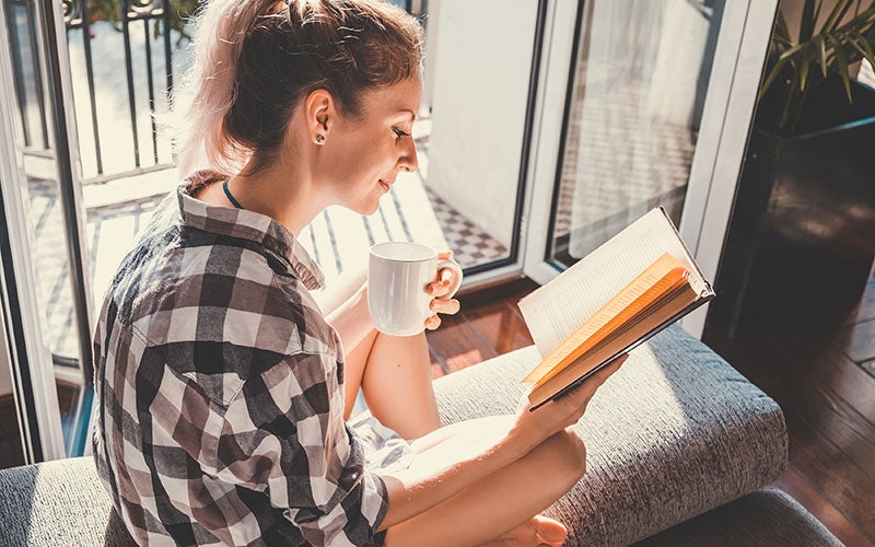 A student reads a book at home while enjoying a cup of coffee.